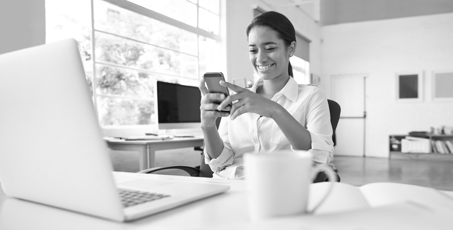 A woman seated at a desk smiles while looking at her smartphone.