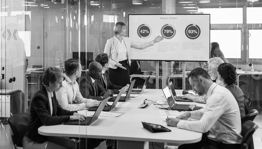 A woman gestures at a PowerPoint presentation in a conference room full of people.