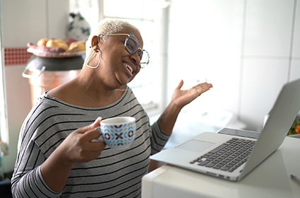 A woman holding a cup of coffee smiles while talking to someone on her laptop.