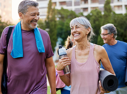 A husband and wife smile at each other as they walk toward an outdoor yoga class.