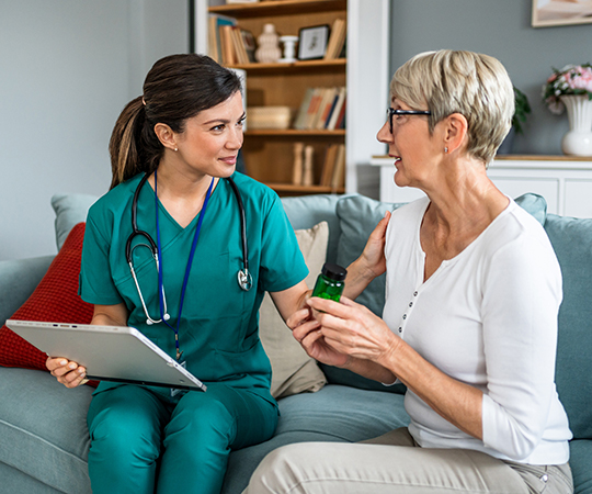 A nurse sits on a couch with a patient in their home while they discuss care options
