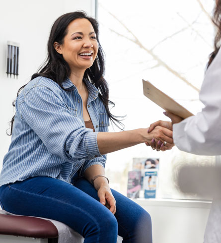 A smiling woman seated on an exam table shaking hands with a health care provider.