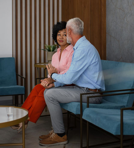 A couple sits together in a waiting room, talking and holding hands.