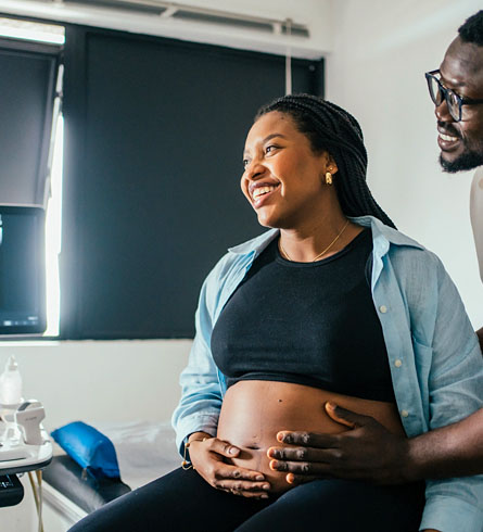A pregnant woman sits in a doctor's office with her partner placing his hand on her stomach.