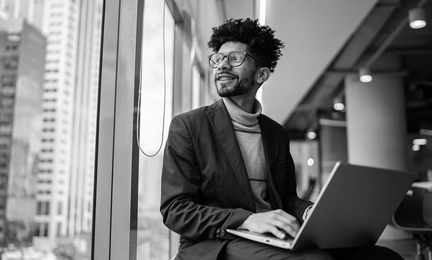 A young man with a laptop looks out an office window 