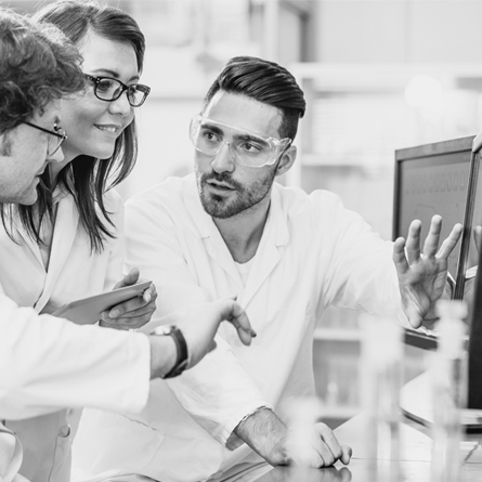 A man in safety goggles explains an experimetn to two people in a lab