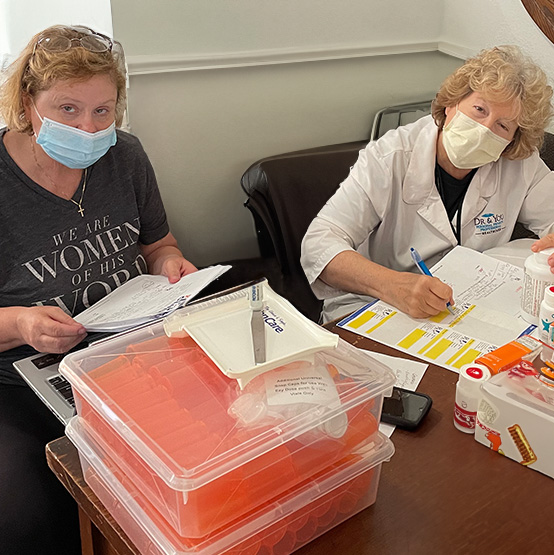 Two clinic staff wearing masks review paperwork at a table with medical supplies.