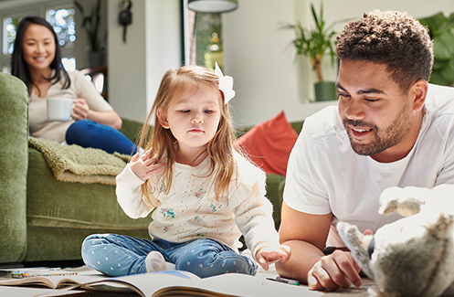 A father and daughter coloring together on the rug, while the mother smiles watching from the couch