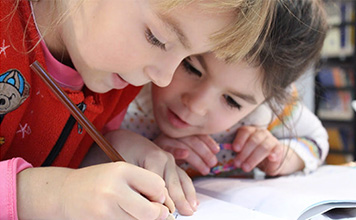 One child writing in a book while the other looks over her shoulder.