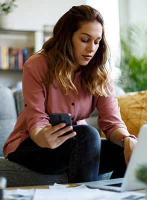 A woman holds her phone in one hand as she types on her laptop