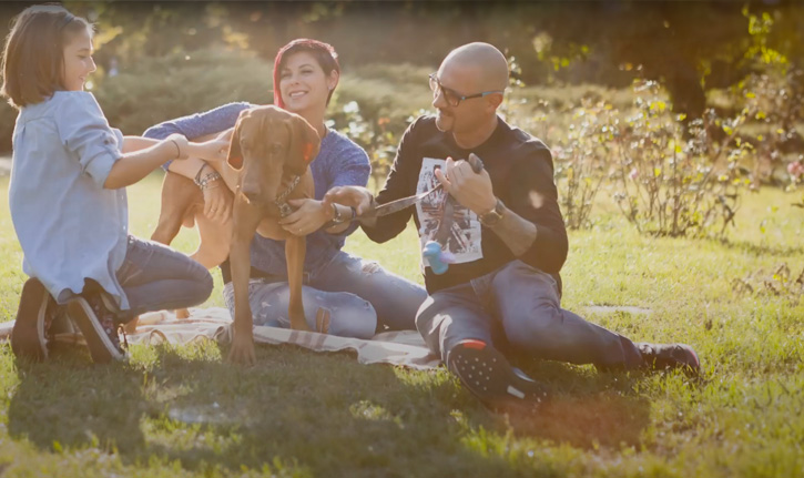A girl and her parents sit on the grass and pet their dog, who is on a leash.