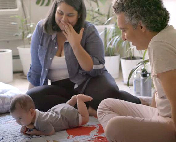 Two adults sit on the floor smiling at a baby lying between them
