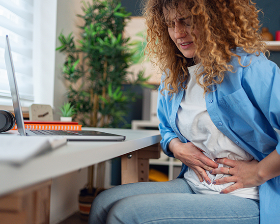 A woman sitting at a desk with a laptop, holding her stomach in pain
