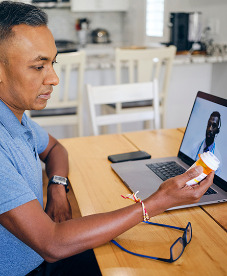 A man sitting at a desk with a laptop, reading a label on a prescription bottle