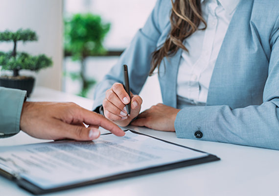 Closeup of a contract on a clipboard with a man's hand pointing toward where to sign as a woman's hand signs her name.