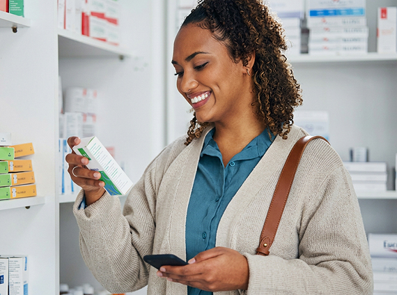 A woman reads a medication label while holding her phone in her opposite hand.