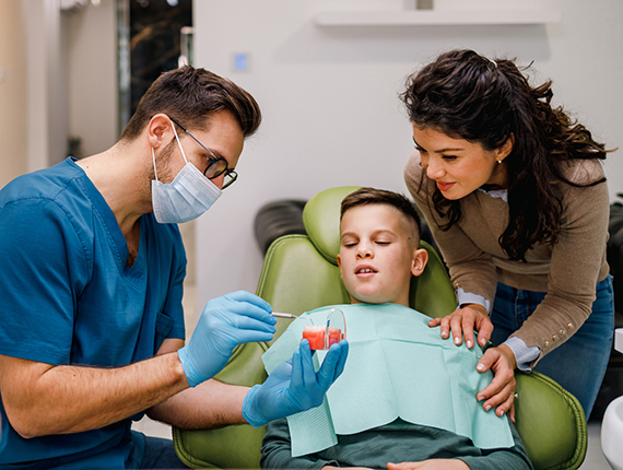 A mother stands over her son in a dentist's chair as the dentist is preparing to do an exam