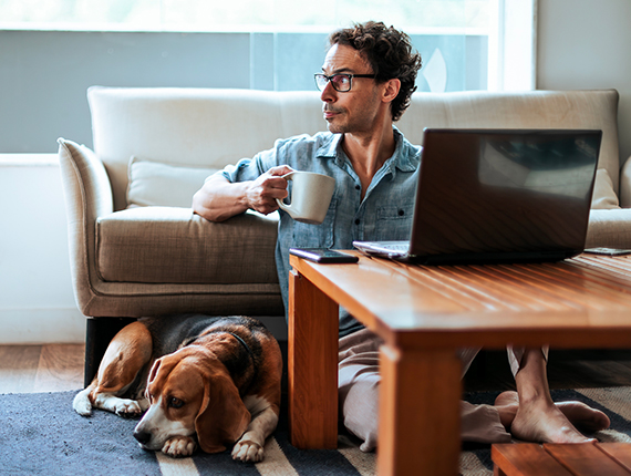 A person sitting on the floor by a laptop on a coffee table, drinking a cup of coffee while a bassett hound is lying on the floor next to him