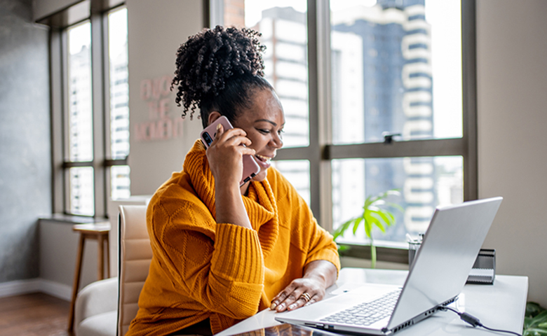 A woman smiles as she talks on the phone in front of her laptop computer.