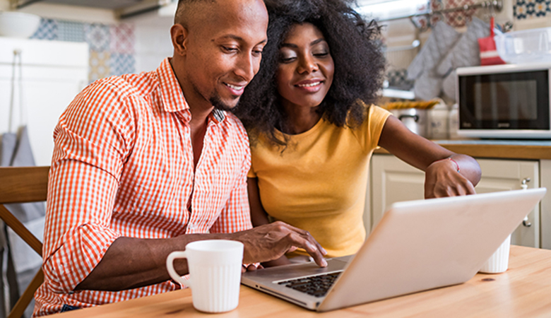 A couple sitting at a kitchen table looks at a laptop together.