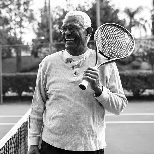 A man laughs as he holds a tennis racket on his shoulder.